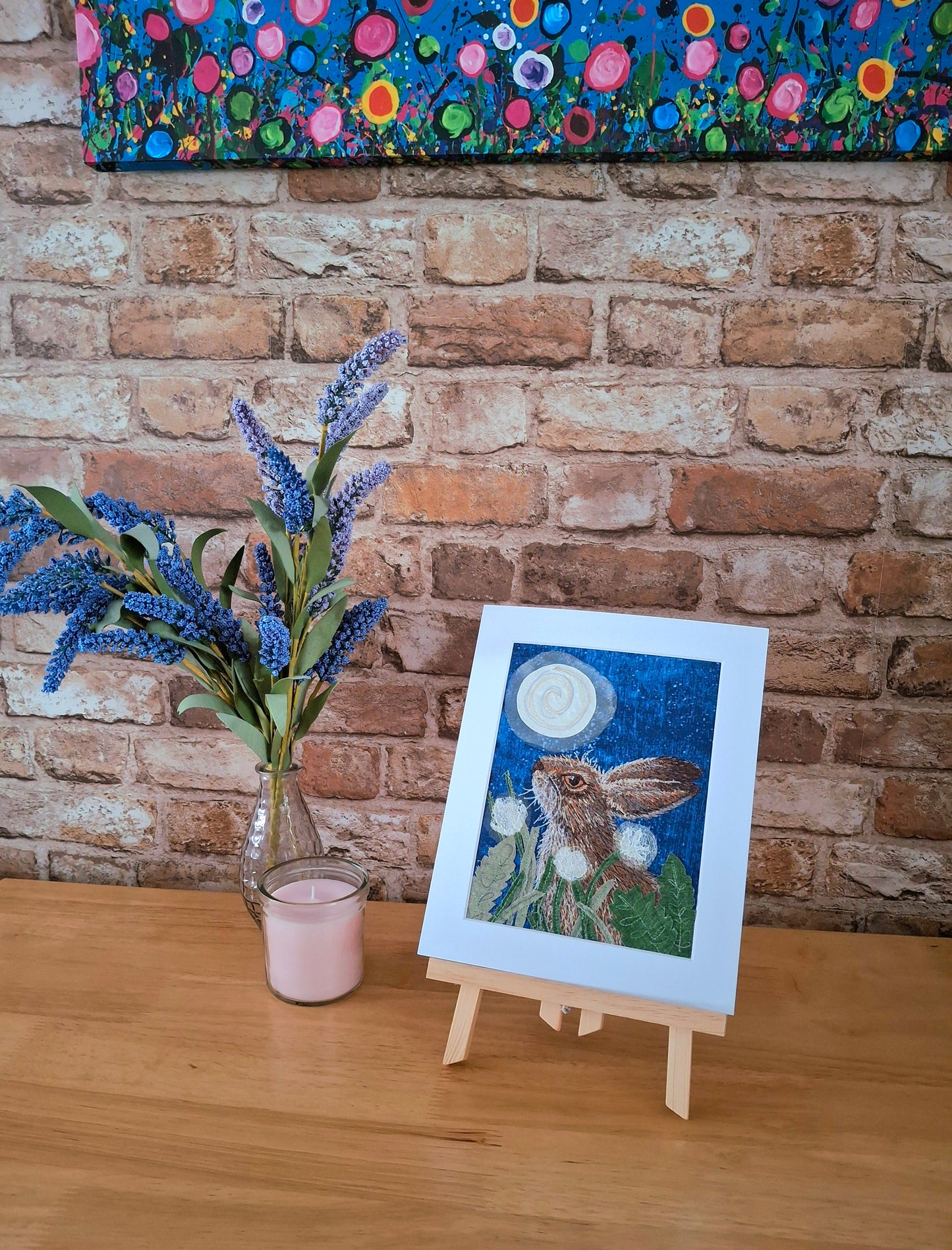 Artwork of a rabbit with flowers on an easel against a brick wall with a colorful floral painting above.