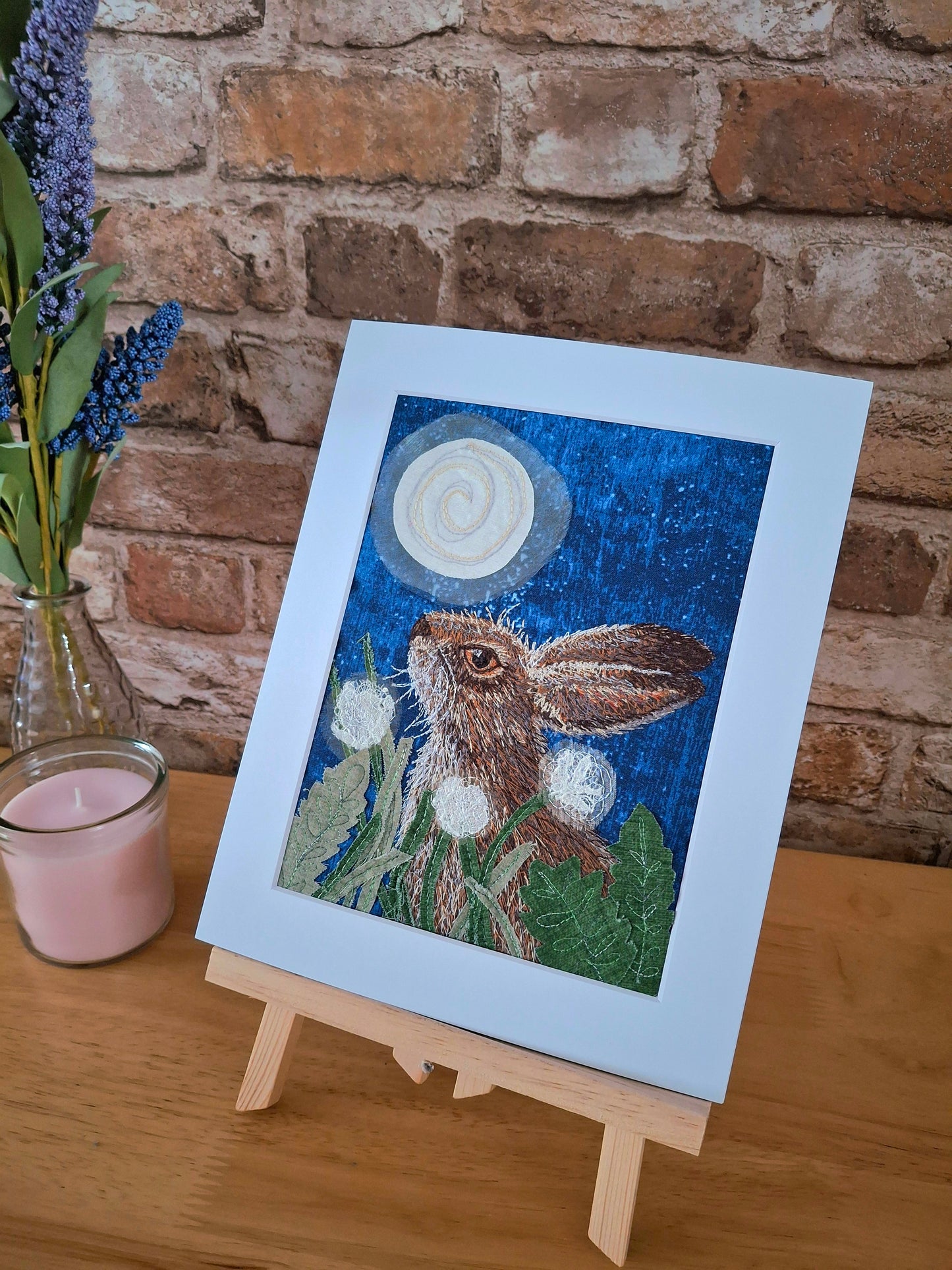 Mounted artwork of a hare with flowers and moon on a wooden stand against a brick wall.