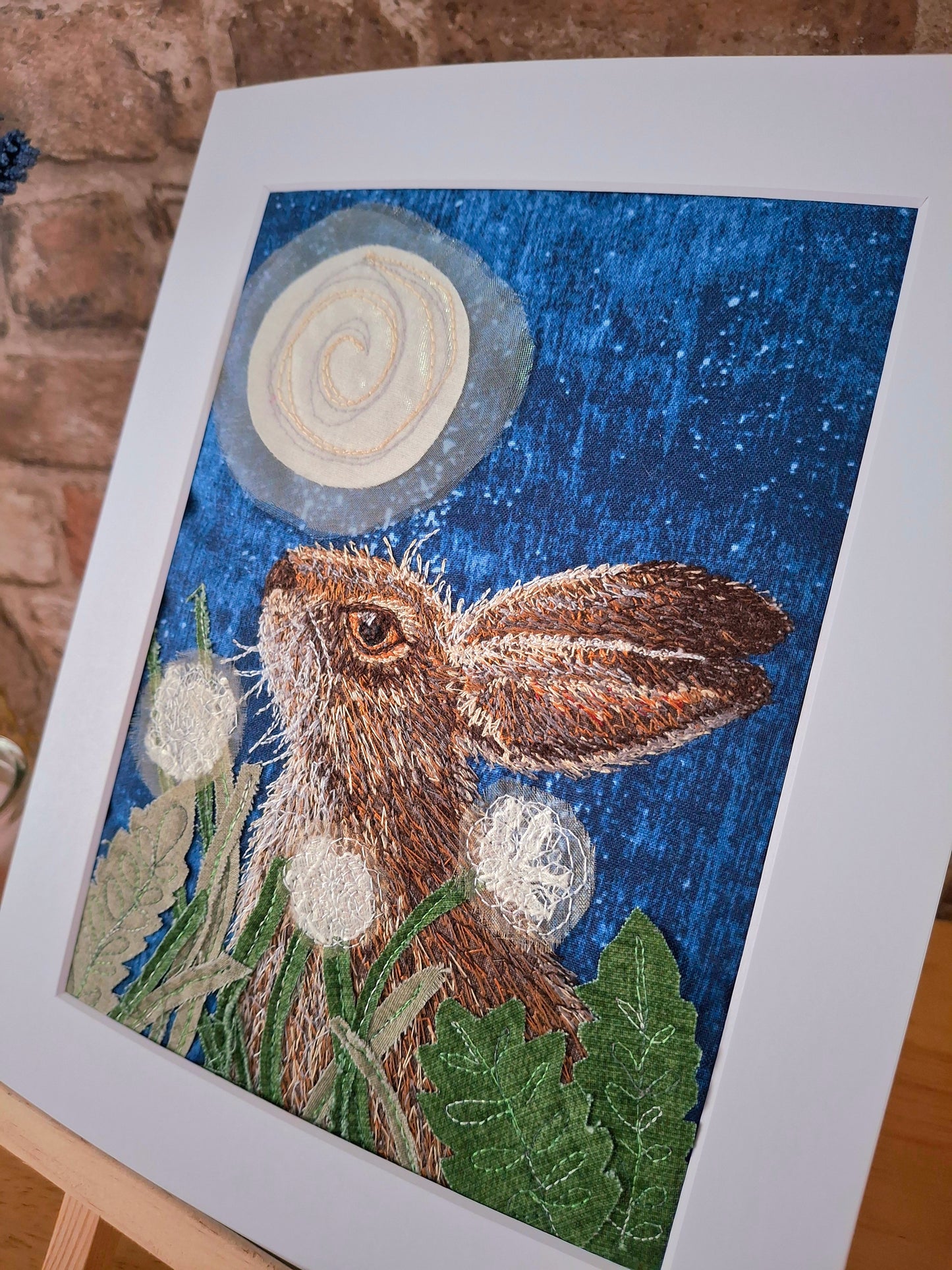 Embroidery of a hare with dandelions and a moon in a white mount.