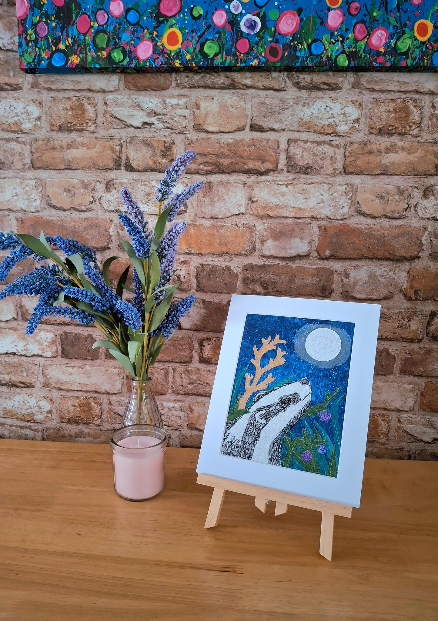 Original artwork of a badger on a small easel with a vase of flowers against a brick wall.