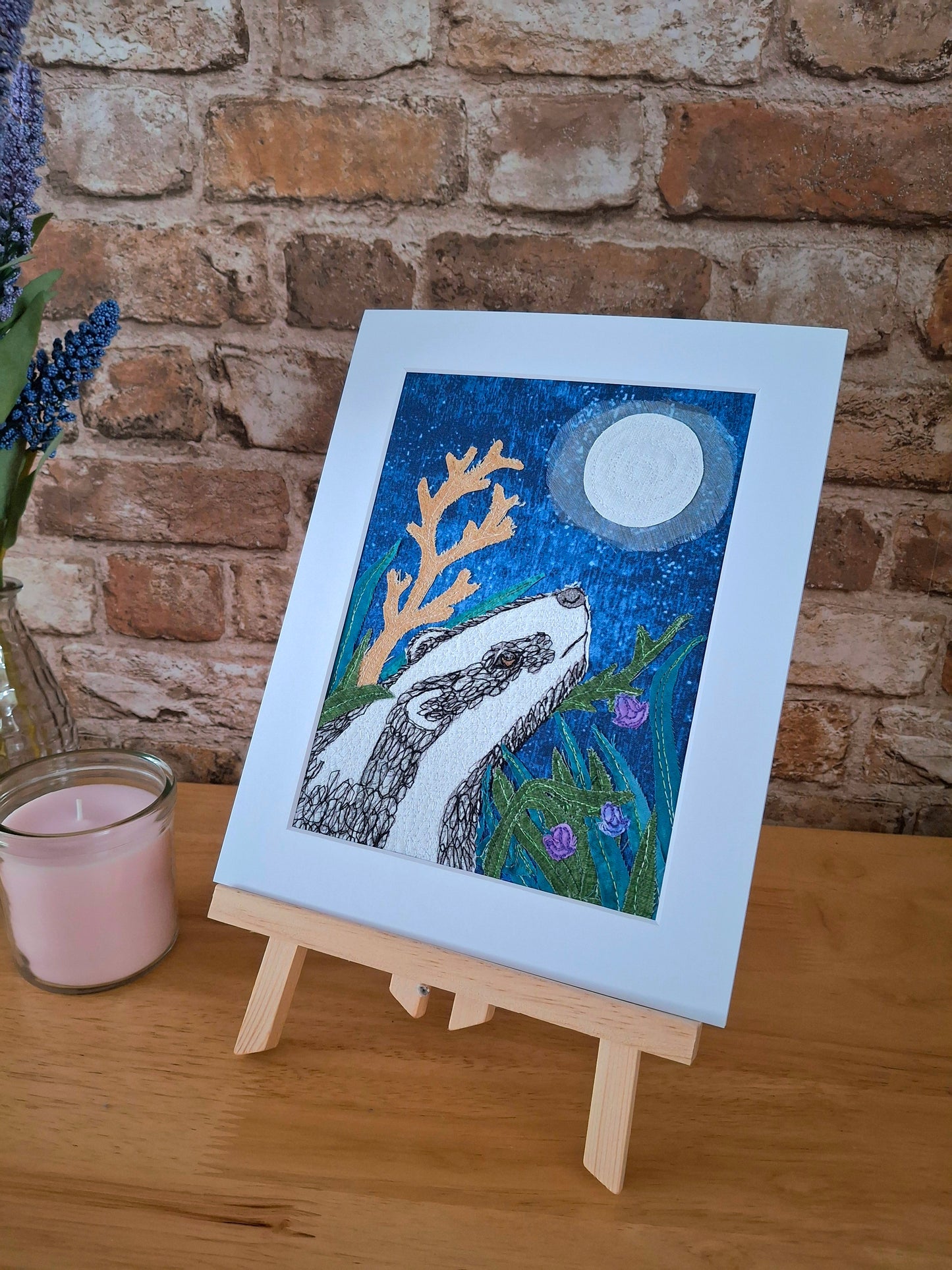 Mounted artwork of a badger with a moonlit sky on a wooden easel against a brick wall.