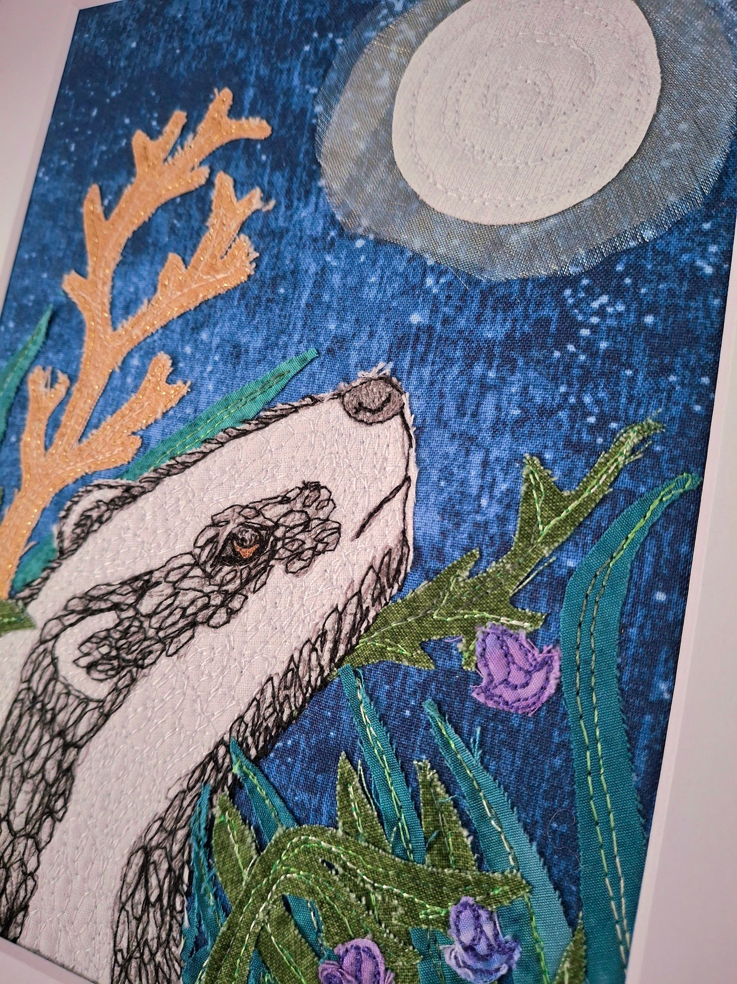 Embroidered artwork of a badger among plants with a moonlit sky.