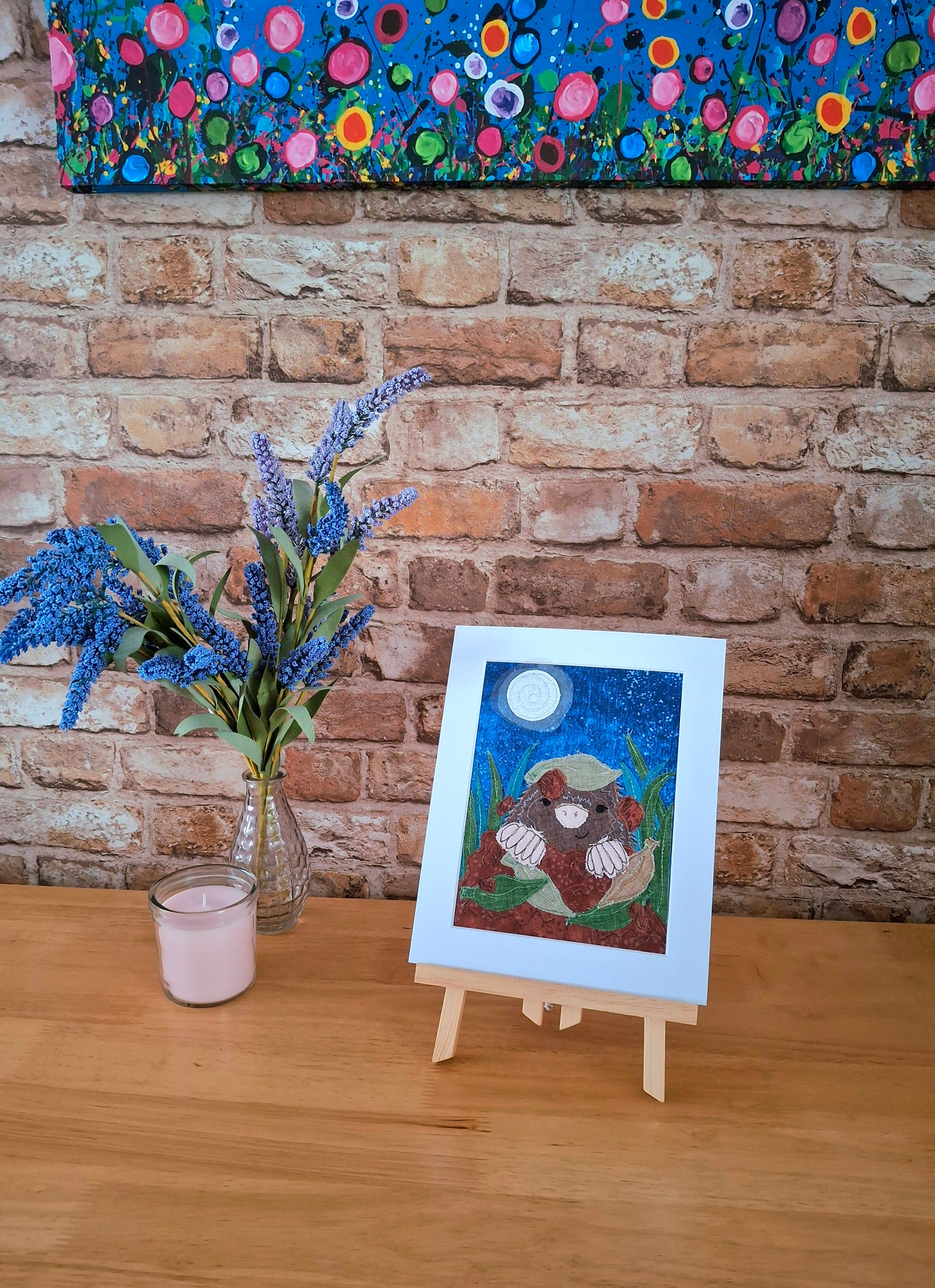 Mole artwork on a wooden easel with flowers and a candle in front of a brick wall and floral painting.