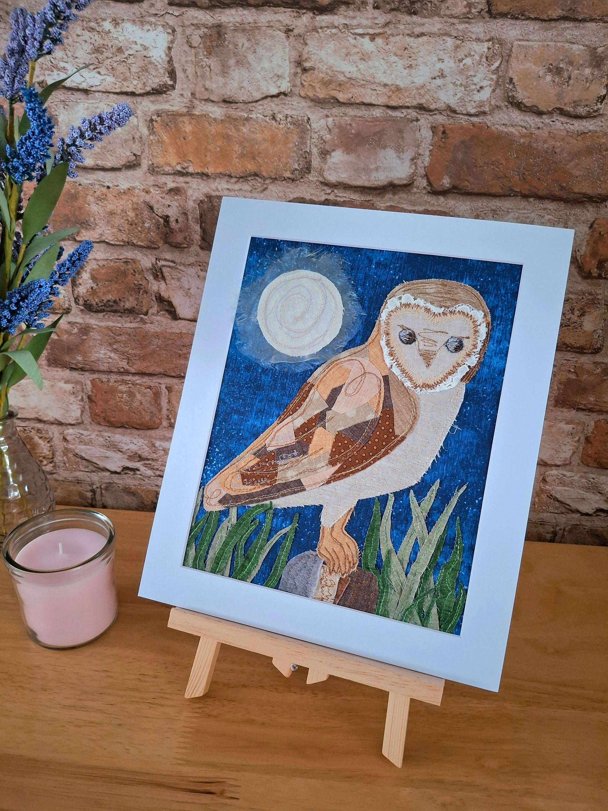 Mounted textile artwork of an owl on a stand against a brick wall with flowers and a candle.